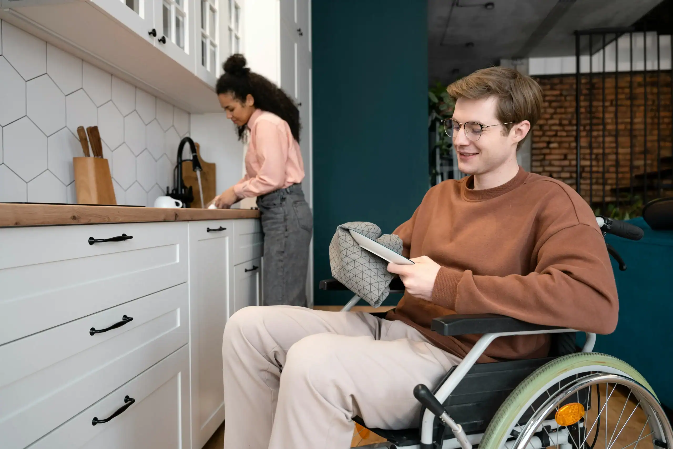 Man in wheelchair smiling in kitchen with caregiver, representing group homes for special needs adults Man in wheelchair smiling in kitchen with caregiver, representing group homes for special needs adults