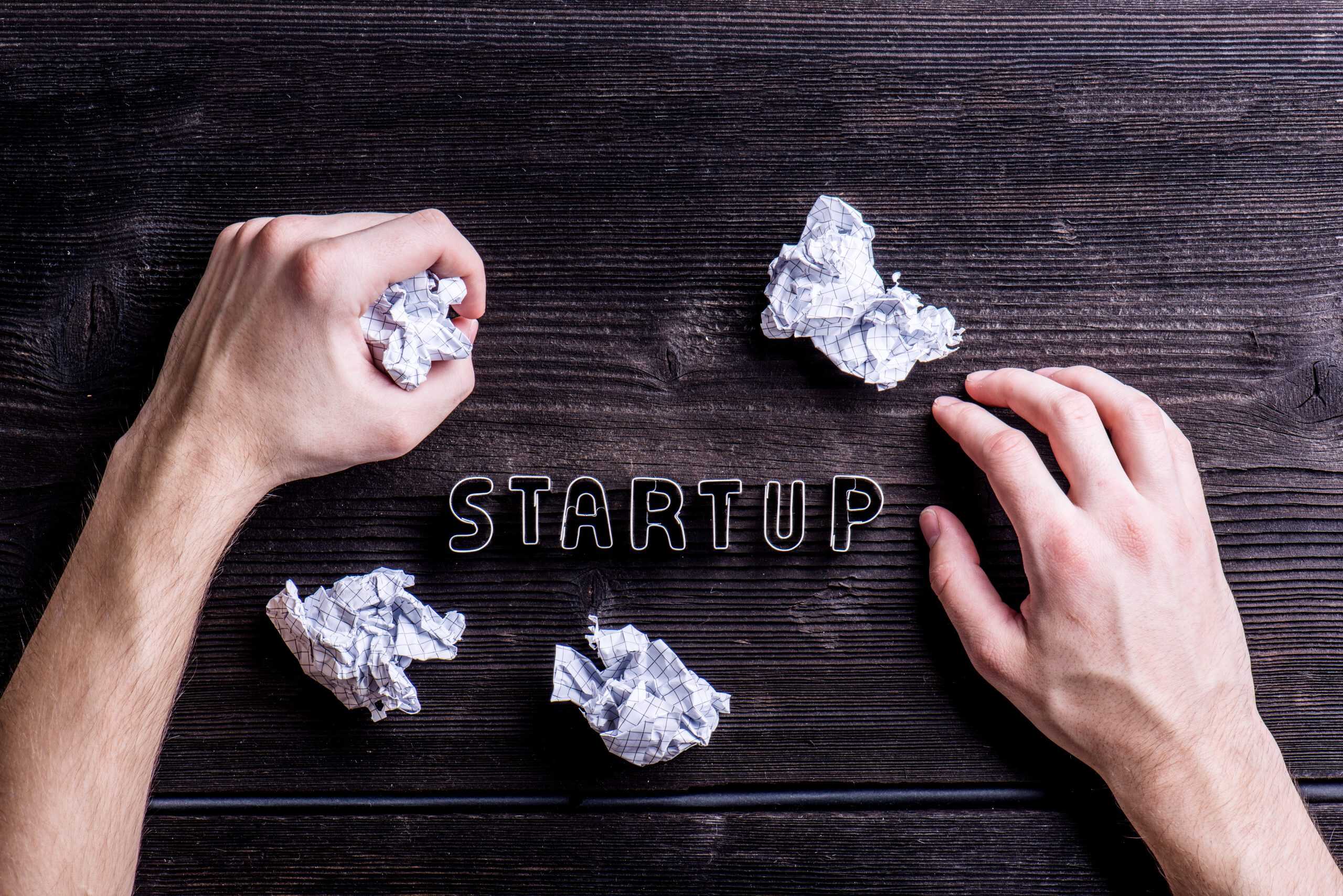 Desk with start up sign, hands of unrecognizable man Office desk with Starting a Small Business Sign made of cookie cutters and hands of unrecognizable man. Crumpled paper balls. Flat lay. Workplace. Studio shot on wooden background.