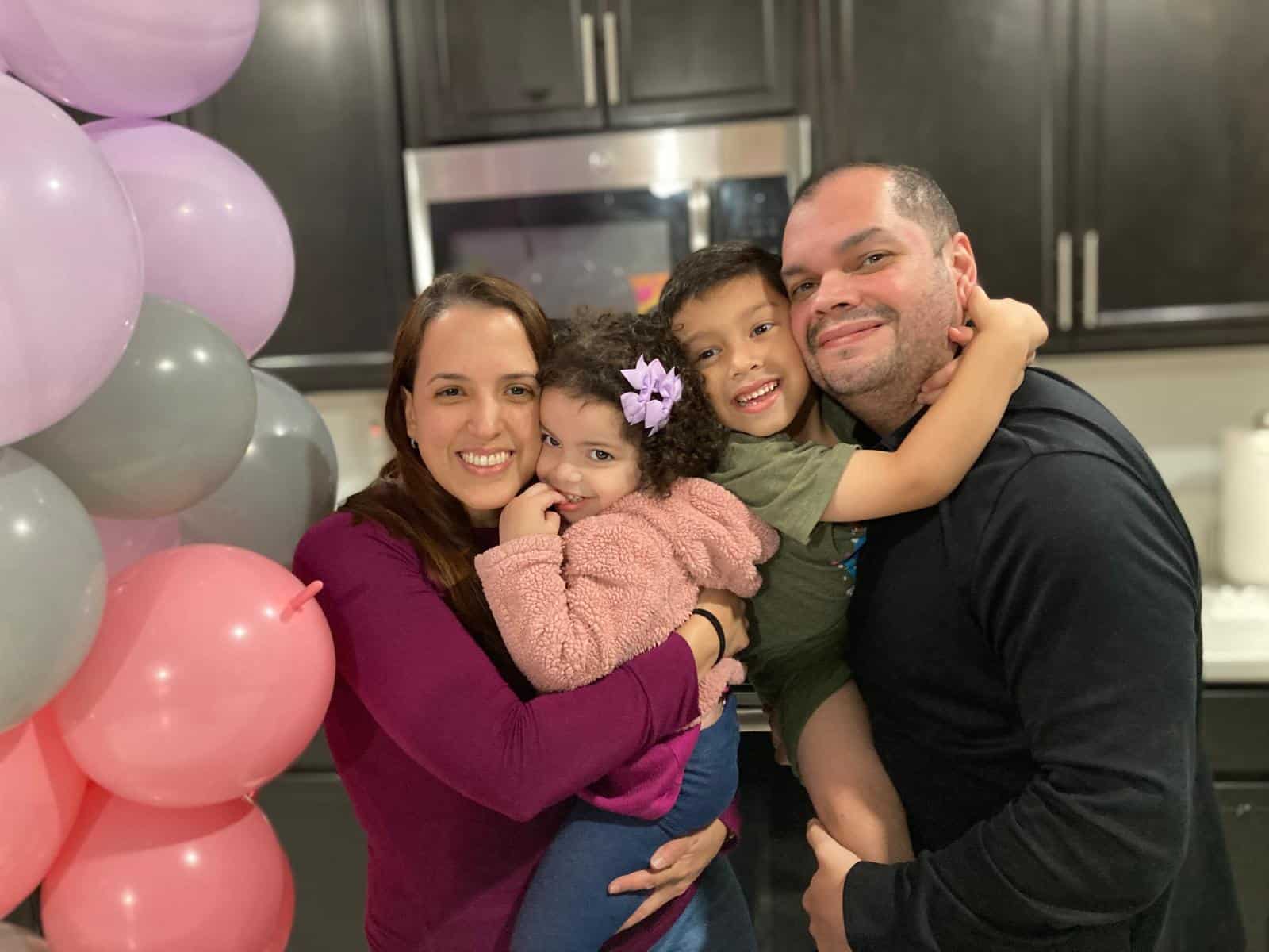 A family of four poses together, smiling. The background features balloons and a kitchen setting.